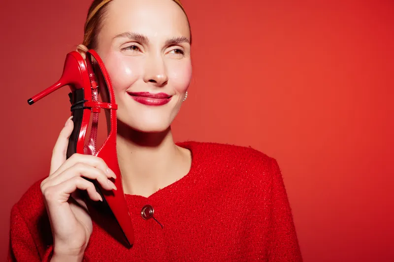 Smiling woman in red sweater holds red high heel shoe near face against red background. Concept of fashion, style, and confident female model with bright makeup and elegant look.