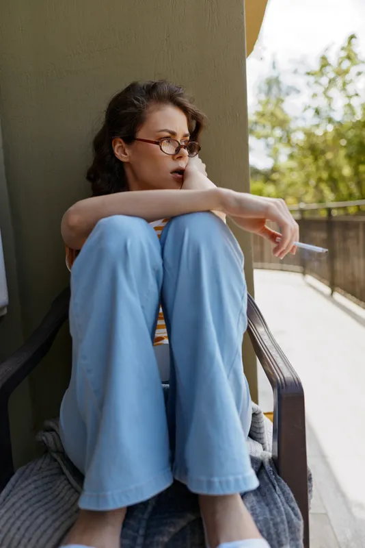 Casual woman sits on a balcony corner, thoughtful mood, wearing light blue jeans and glasses, natural daylight creates a calm intimate moment at home, soft shadows present.