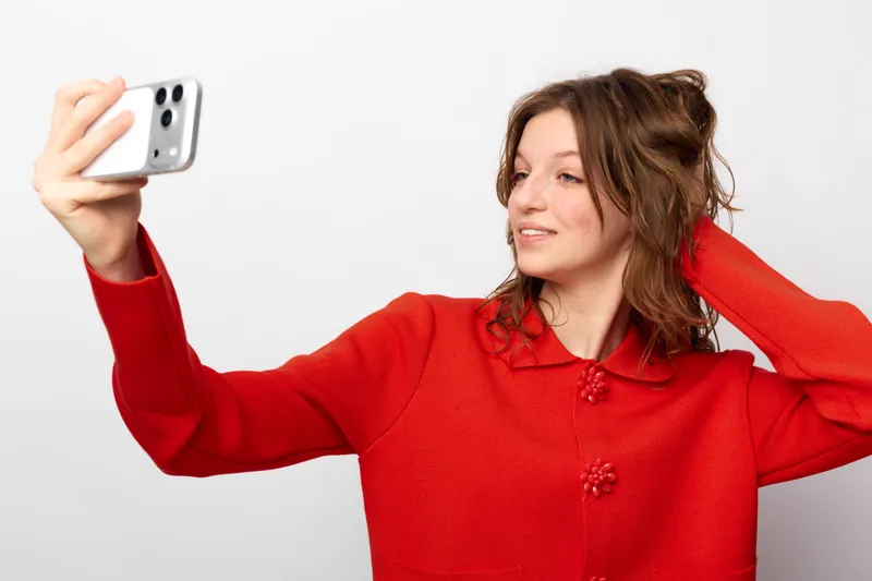 Young woman in red sweater takes a selfie with smartphone, smiling and posing against a plain white background. Concept of social media, self-portrait, and casual lifestyle.