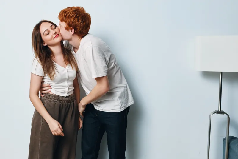 Young couple in casual clothes showing affection with a kiss on the cheek in a bright modern interior with lamp. Romantic moment with love and happiness between man and woman.