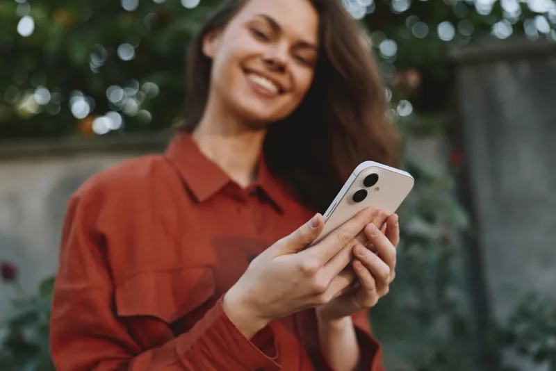 Smiling woman with shoulder length hair wearing rust shirt looking at a phone outdoors. Natural light highlights a relaxed lifestyle moment with casual fashion and everyday connectivity, emphasizing