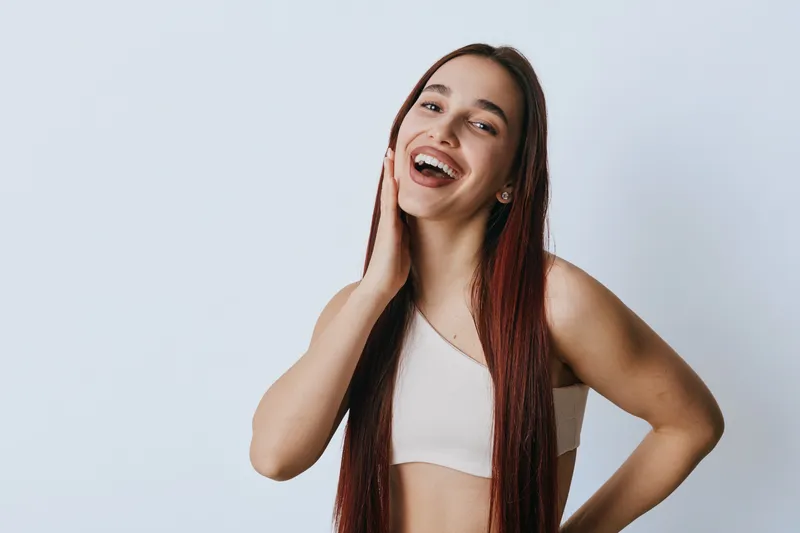 Young woman with long hair smiling and touching face in casual top against plain background. Happy female portrait with natural expression and relaxed pose for lifestyle or beauty concept.