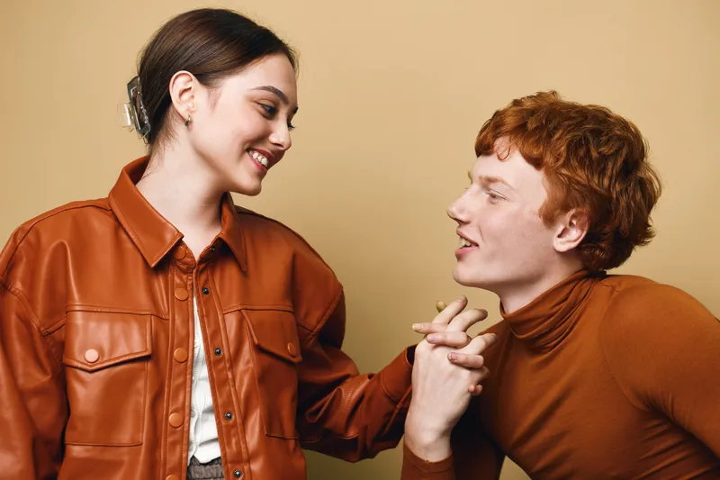Couple, woman and man holding hands, smiling and looking at each other against beige background in casual cozy autumn clothes, friendship and connection.