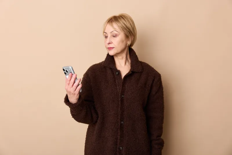Woman with short blonde hair holds a smartphone in her hand, wearing a dark coat. Portrait against a beige studio background, suggesting communication and messaging.