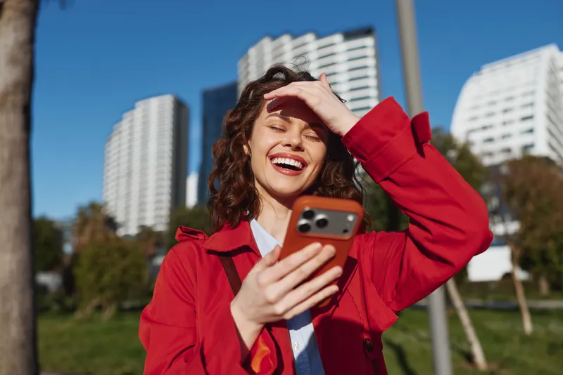 Smiling woman with curly hair in a bright red jacket holds a smartphone outdoors in an urban park with modern buildings in the background, enjoying a sunny day.
