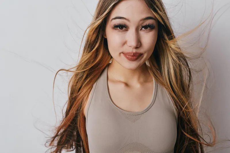 Young woman standing with long hair blowing in the wind wearing sleeveless top looking confidently at camera smiling softly indoors with neutral background.