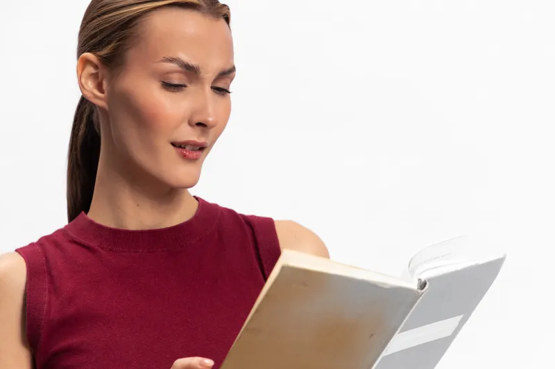 Young woman in red sleeveless top reading a book with focused expression. Isolated on white background. Concept of education, learning, concentration, and knowledge acquisition.