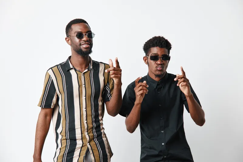 Two African American men with dark skin wearing sunglasses and casual shirts posing and gesturing with fingers on white background. Young black males with confident expressions.