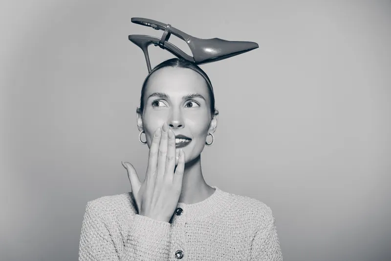 Surprised Caucasian woman with pale skin and short hair holding hand near mouth and wearing high heel shoe on head in studio.