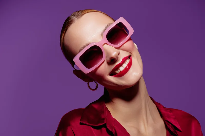 Smiling woman with pink sunglasses and red lipstick poses against purple background. Fashion portrait with bright colors and confident expression. Concept of style and beauty.