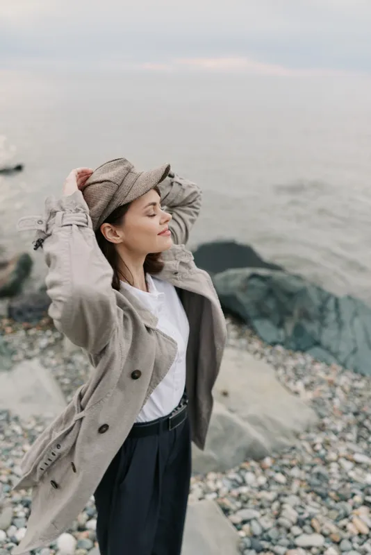 Young woman wearing beige coat and cap enjoys fresh sea breeze with closed eyes on rocky beach. Concept of relaxation, freedom, nature, and peaceful outdoor moment by the ocean.