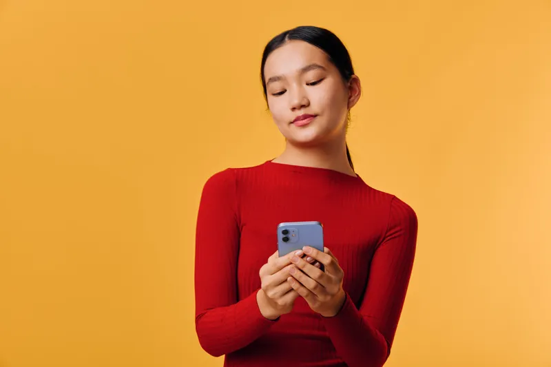 Young woman in red sweater holding smartphone looking down at device with slight smile on yellow background. Casual portrait of female using mobile phone for communication or social media.