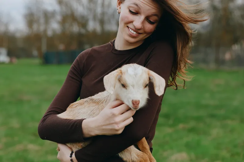 Young woman with long hair smiles gently while holding a baby goat outdoors in a casual brown sweater during a relaxed lifestyle moment in nature.
