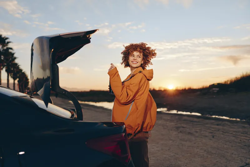 Woman with curly hair near open car trunk on beach at sunset, golden light, casual jacket, standing outdoors, road trip lifestyle, ocean waves behind, relaxed smile