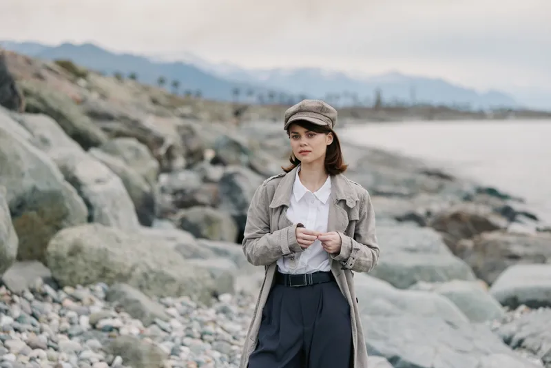 Young woman in stylish coat and hat stands on rocky beach near calm sea. Overcast sky and distant mountains create serene, contemplative mood for fashion and nature concept.