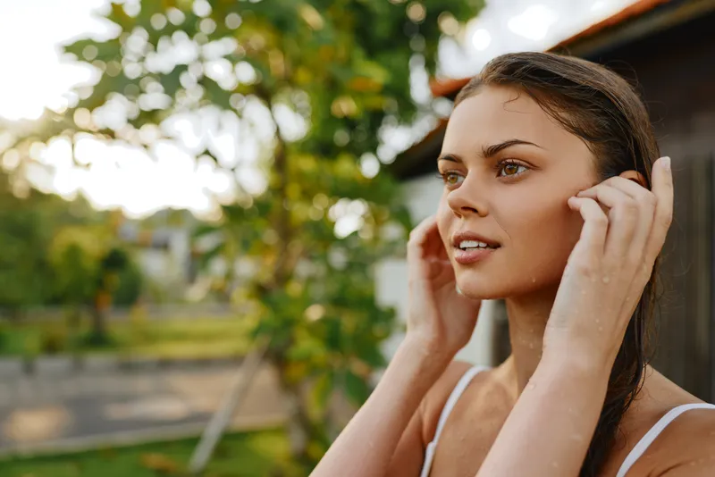 Natural outdoor portrait of a woman adjusting her hair, casual summer style, calm confident mood, sunlit garden setting, everyday beauty and candid lifestyle moment captured.