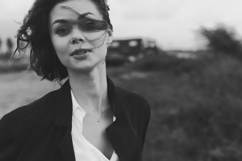 Woman in a monochrome outdoor scene with windy hair, wearing a blazer and white shirt, exudes fashion mood in candid street style photography, conveying confidence and urban atmosphere