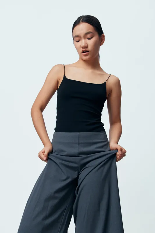 Young woman wearing black tank top and loose gray pants stands with hands in pockets against plain background showing confident casual pose and relaxed expression in studio lighting.