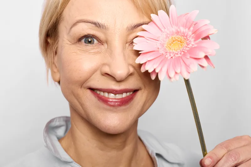 Smiling woman holding a pink gerbera flower near her eye, soft studio lighting on light background, concept of skincare wellness self care, close up portrait beauty