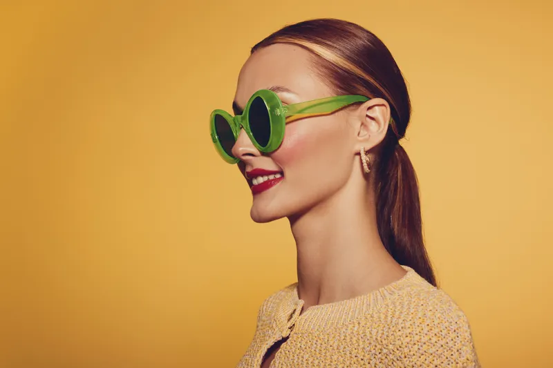 Young woman with green sunglasses and red lipstick smiles confidently against yellow background. Fashion portrait with bright colors and stylish accessories in studio lighting.