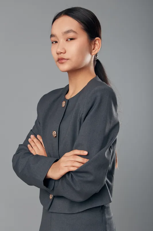 Confident young woman standing with crossed arms wearing formal dark grey business outfit against grey background. Portrait of female with long black hair and serious expression in studio.