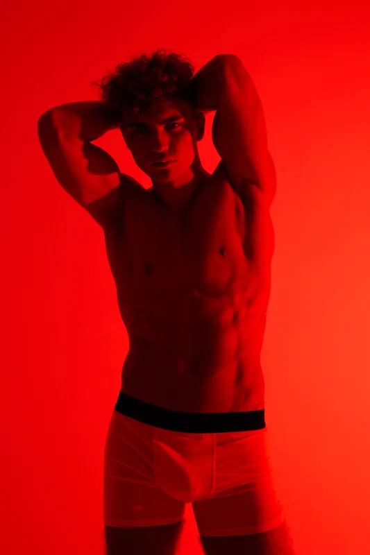 Man posing in white underwear on red background with muscular body and curly short hair showcasing fitness and modern fashion in studio lighting for health and lifestyle.