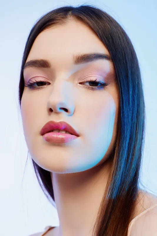 Close-up portrait of a woman with long dark hair and glossy pink lips. Soft studio lighting highlights her smooth skin and subtle makeup. Concept of beauty, fashion, and skincare.