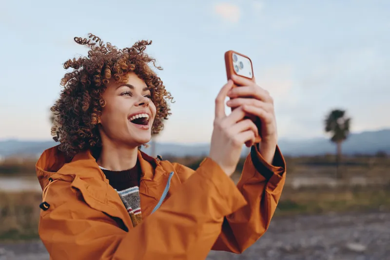 Smiling woman in orange jacket takes a smartphone selfie outdoors in daylight, curly hair blowing, casual lifestyle portrait against mountains and open sky, natural vibe for travel freedom