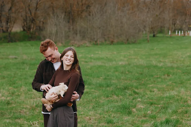 A young couple embraces outdoors as the woman holds a small baby goat in her arms, sharing a joyful moment in a green field during early spring.