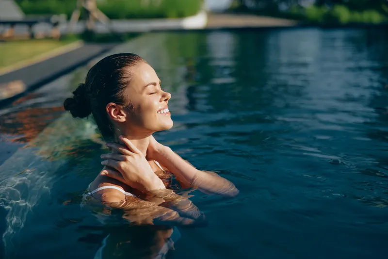 Cheerful woman in a sunlit pool, eyes closed, smiling softly, enjoying a relaxing moment outdoors with warm breeze, clear water and natural colors, serene mood, carefree summertime vibe