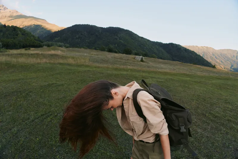 Young woman outdoors bends forward in a grassy plain with a backpack, enjoying freedom and fresh air in a remote hillside landscape