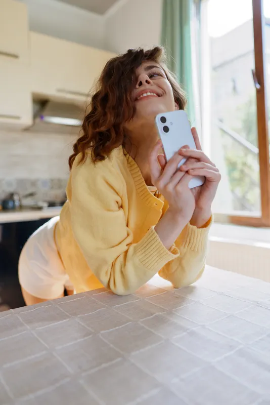 Cheerful woman in a yellow sweater leans over a kitchen table, holding a smartphone and smiling at the screen, capturing a casual home moment with warm, inviting vibe and lifestyle emphasis
