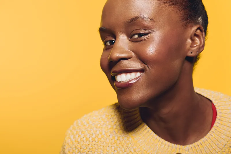Smiling African American woman with dark skin wears yellow sweater against orange background. Close-up portrait shows natural beauty, happiness, and confidence in warm studio lighting.
