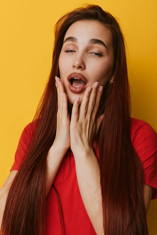Young woman with long red hair and red shirt expressing surprise or shock with open mouth and hands on cheeks against yellow background. Emotion, face and reaction closeup.