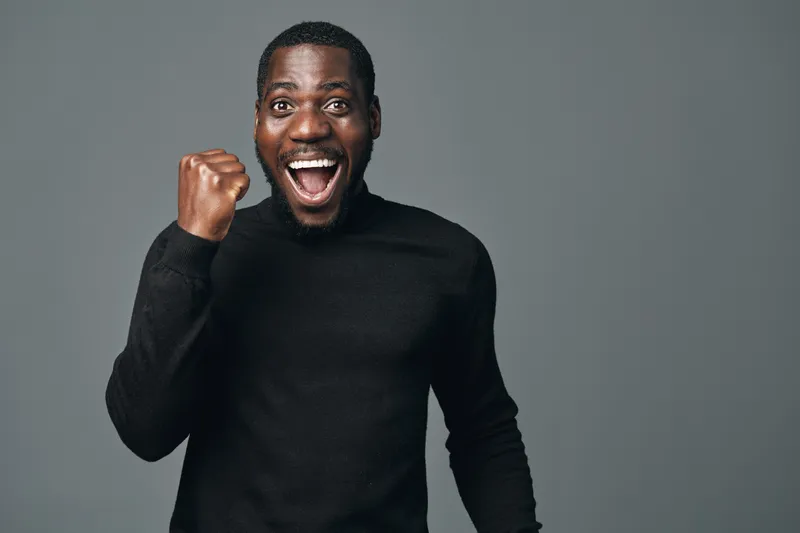 African man with dark skin wearing black sweater celebrating success with fist pump and wide smile in studio. Creative conceptual visual style with happy expression and positive energy.