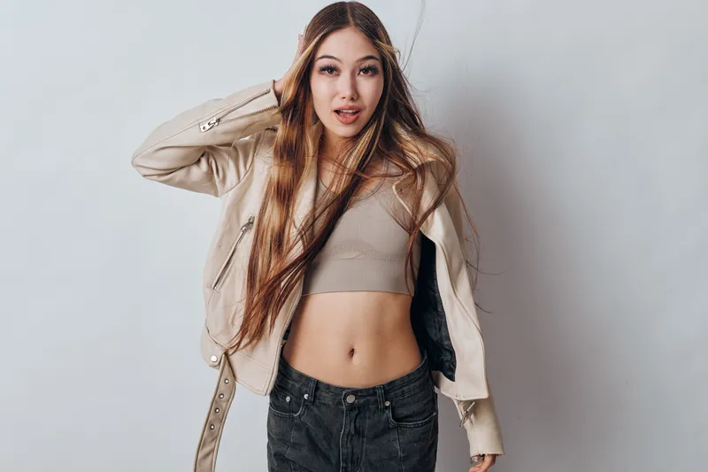 Young woman with long hair wearing crop top and jacket posing, looking confident against light background with casual style and fashion outfit for youth.