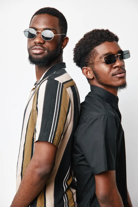 Two African American men with dark skin wearing sunglasses and casual shirts standing back to back against white background. Stylish young male models with confident poses.