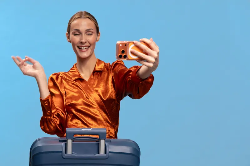 Smiling woman in orange satin blouse takes selfie with smartphone while standing behind blue suitcase on blue background. Concept of travel, vacation, lifestyle, and happiness.