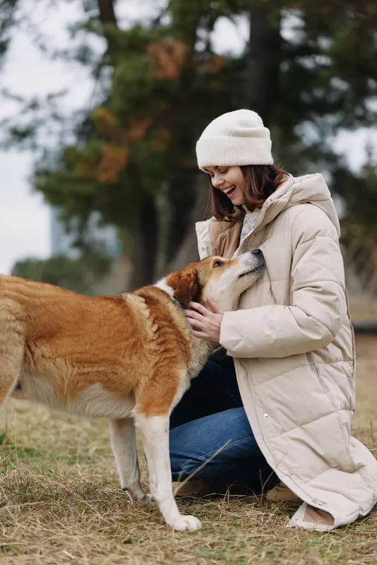 Woman in a warm coat and beanie kneels by a park path, smiling while gently petting a friendly dog beside autumn grass and trees on a calm, breezy day in view.
