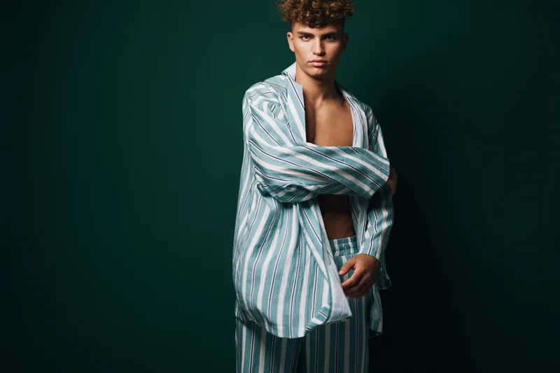Young man with curly hair wearing striped pajamas posing with crossed arms against dark background. Fashion, casual style and confident expression in studio portrait.