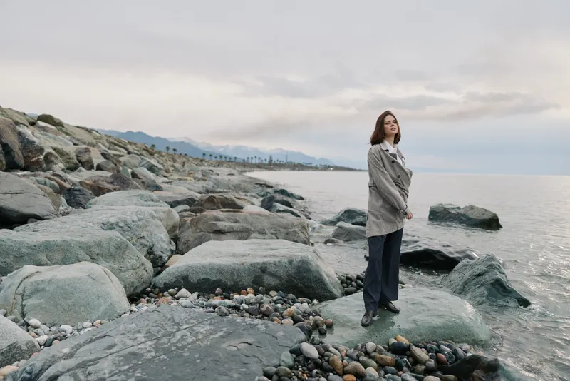 Young woman in casual clothing stands on rocky beach near calm sea under cloudy sky. Natural landscape with stones and distant mountains creates peaceful and contemplative mood.
