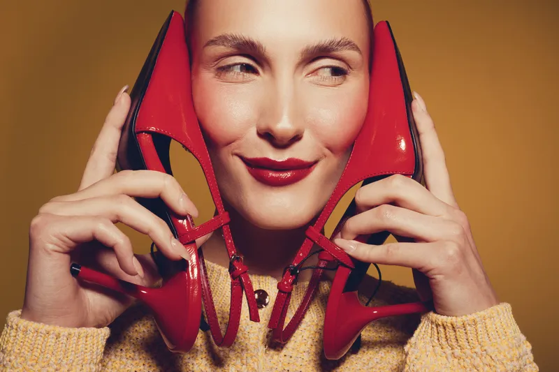 Smiling woman holds red high heel shoes near face, wearing yellow sweater. Studio portrait on warm brown background. Concept of fashion, style, and playful mood.