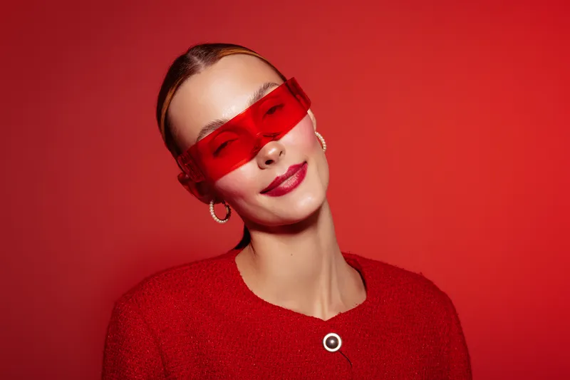 Young woman in red sweater and red sunglasses poses smiling on red background. Fashion portrait with vibrant colors and confident expression. Concept of style and modern look.