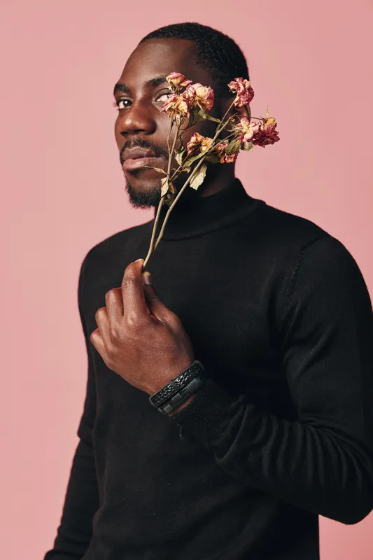 African man in black turtleneck holding dried flowers near face with creative conceptual visual style on pink background. Portrait with thoughtful expression and artistic mood.