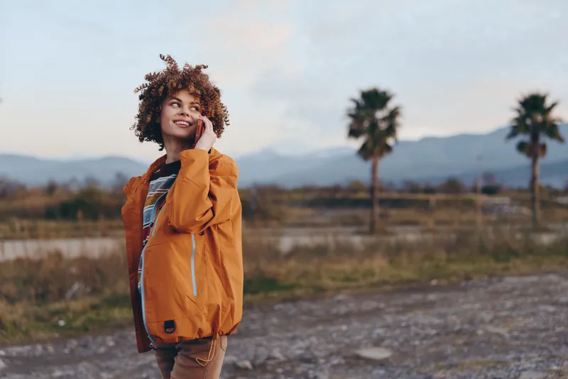 Woman lifestyle outdoors, smile in orange jacket, curly hair in soft wind, standing near palm trees, distant mountains behind, golden hour light, natural colors
