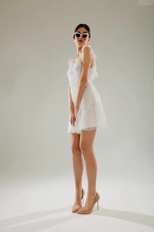 Fashionable woman in a white textured dress poses confidently wearing stylish sunglasses and nude high heels. Studio shot on a neutral background. Concept of beauty, fashion, and elegance.