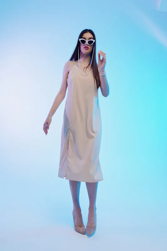 Tall woman in a beige dress and white sunglasses poses confidently against a blue gradient background. Concept of woman beauty fashion and modern style.