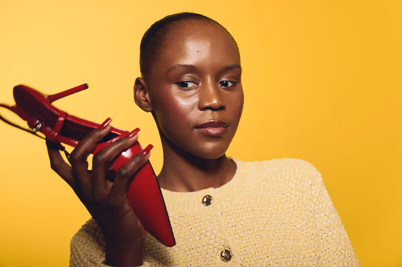 Portrait of a confident black woman with dark skin holding a red high heel shoe near her face. Yellow background, fashion concept, style, elegance, and beauty in studio lighting.