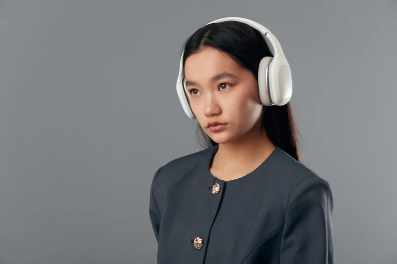 Young woman wearing white headphones and gray jacket standing against gray background looking thoughtful and focused. Listening to music or audio with modern wireless headset indoors.