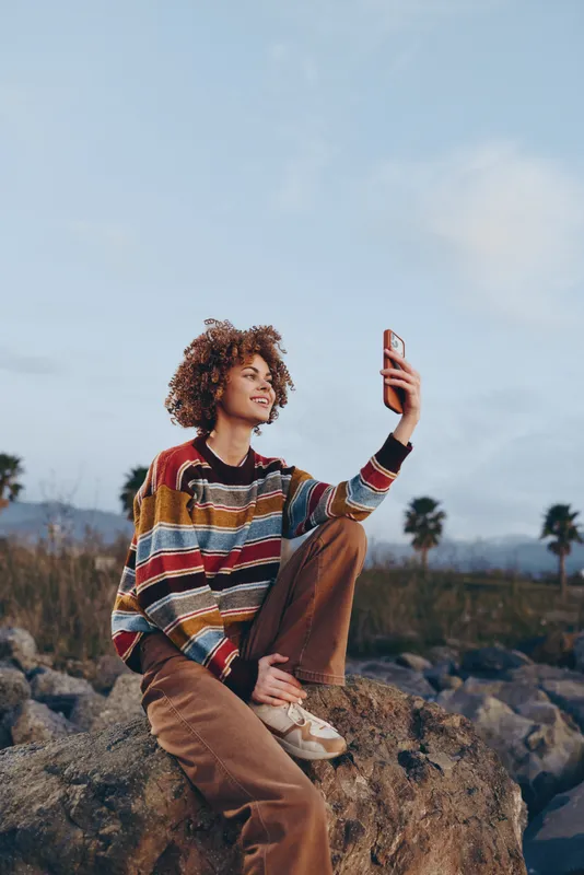 Woman selfie smartphone lifestyle outdoors rock curly hair striped sweater casual pose on stone, smiling and looking at camera, warm daylight, mountain landscape with palm trees horizon.